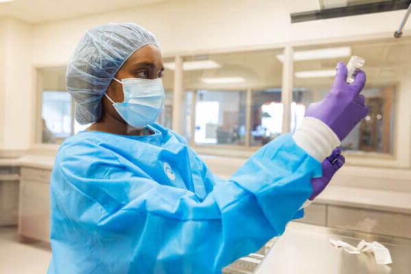 全球最大的博彩平台. Dunn School of Pharmacy student in blue scrubs, gloves, cap and mask, holding a vial.